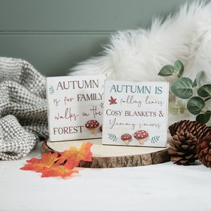 Two Autumn Easel Signs With 3D Resin Toadstools are displayed on a wooden slice with pinecones, autumn leaves, and 3D resin toadstools. One sign reads "Autumn is for family walks in the forest," and the other says "Autumn is falling leaves, cosy blankets & yummy s'mores." A cozy blanket is in the background.