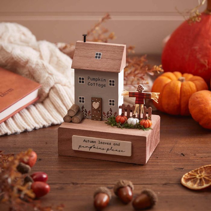 A small wooden decoration labeled "Autumn Pumpkin 3D House With Scarecrow Scene" sits on a table surrounded by autumn-themed items. The whimsical Scarecrow Scene includes pumpkins, acorns, leaves, and a small scarecrow. A sign on the base reads, "Autumn leaves and pumpkin please.