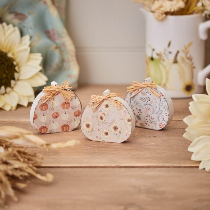 Three decorative pumpkins made of fabric, each with different pastel patterns, are arranged on a wooden surface. The Autumn Rustic Design Wooden Pumpkins With Raffia Bow are surrounded by dried flowers, white sunflowers, and a ceramic jug with a pumpkin design.