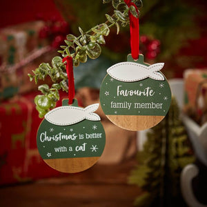 Two Christmas ornaments hang from a leafy branch. The ornament on the left reads, "Christmas is better with a cat," in white text on a green background—the perfect cat lover gift. It features the *Christmas Cat Hanger Green Wood With 3D Fish.* The ornament on the right reads, "Favourite family member," in white text on a green background.