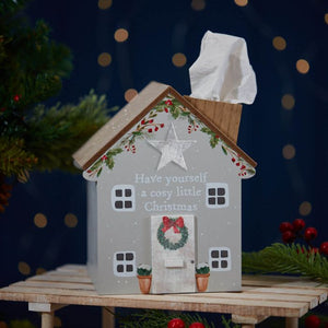 A Christmas Cosy House Tissue Box Holder adorned with festive Christmas decor is placed on a small table. The holder is designed to look like a house, featuring a wreath on the door, small plants flanking the entrance, and garlands adorning the roof. Tissues are dispensed through the chimney. The text reads, "Have yourself a cosy little Christmas.