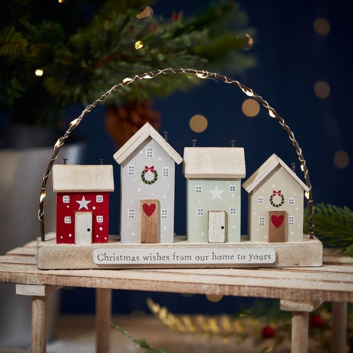 The "Christmas Family Wishes Block Wooden Houses With LED Wire Arch" features four wooden houses adorned with wreaths and stars, arranged on a wooden base inscribed with "Christmas wishes from our home to yours." The background showcases a blurred view of a Christmas tree with twinkling lights, creating warm family wishes.