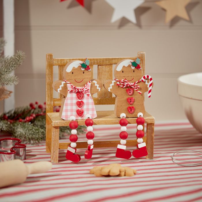 A decorative scene featuring the Christmas Gingerbread Girl & Boy Shelf Sitters perched on a small wooden bench. The figures, dressed with icing and red bead legs, are surrounded by festive decor such as a rolling pin, cookie cutters, and star decorations. Striped fabric covers the surface below.