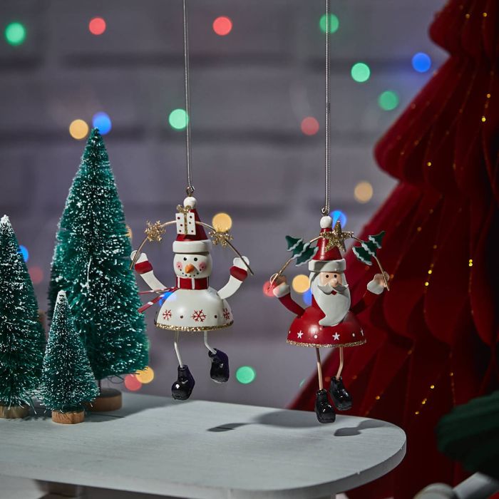 Two festive Christmas Metal Springy Snowman & Santa Hangers, shaped like a snowman and Santa Claus, are hanging. Both hold star wands and wear festive attire. Surrounded by small, decorated Christmas trees with multi-colored lights blurred in the background, these ornaments evoke a sense of festive magic.