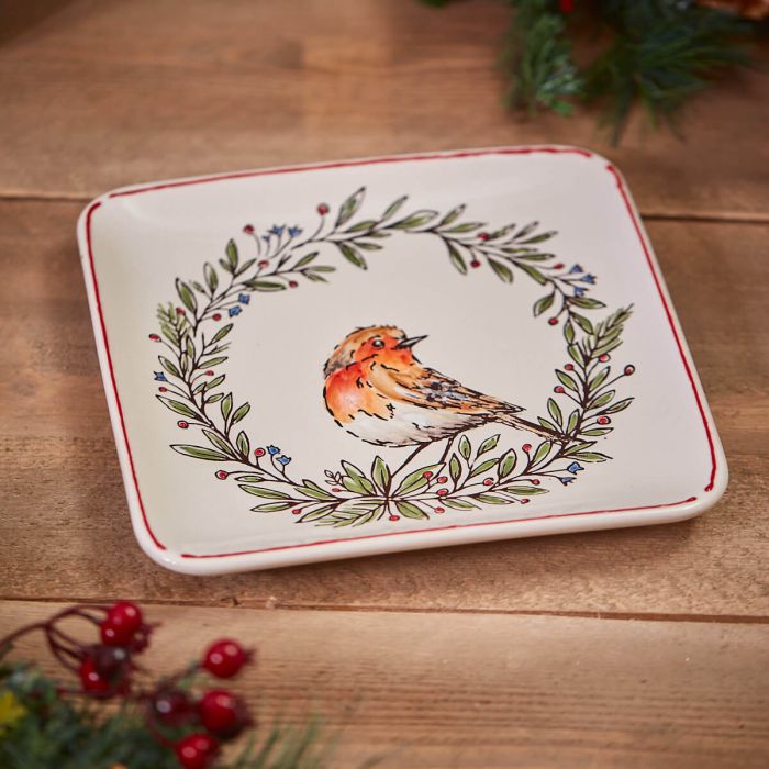 The Holiday Tableware collection now features the Christmas Robin & Wreath Square Stoneware Dish. This ceramic plate has a red rim and showcases a painted image of an orange and brown bird in the center, encircled by a wreath of green leaves and blue berries. It's set on a wooden surface with some red berries and greenery nearby for added festive charm.