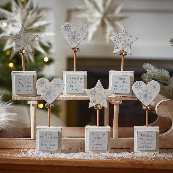 A festive display of Christmas Star & Heart Block With Quote, each adorned with heart block accents and star-topped stands featuring various Christmas messages. These messages include classic holiday wishes and lyrics. The background showcases a beautifully decorated Christmas tree with soft lights and snowy decor, creating a warm atmosphere perfect for the holiday season.