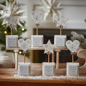 A festive display of Christmas Star & Heart Block With Quote, each adorned with heart block accents and star-topped stands featuring various Christmas messages. These messages include classic holiday wishes and lyrics. The background showcases a beautifully decorated Christmas tree with soft lights and snowy decor, creating a warm atmosphere perfect for the holiday season.