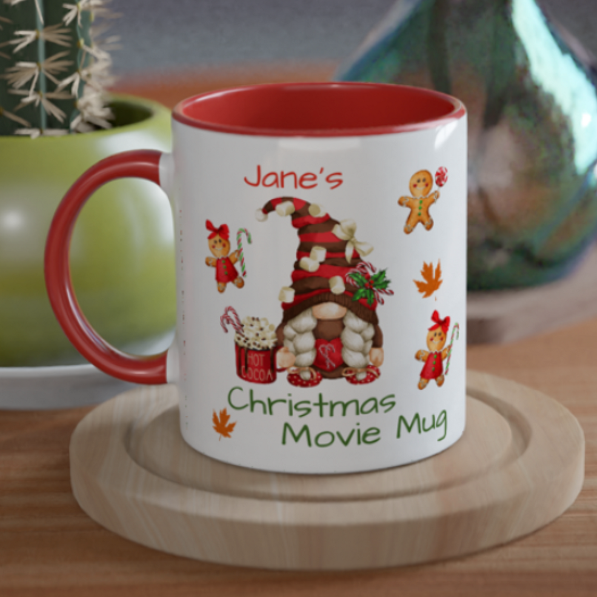 A festive mug sits on a wooden surface. With a red interior and a holiday design featuring a gnome house made of marshmallows, gingerbread figures, and candy canes, it reads "Personalised Christmas Movie Mug." This personalised Christmas mug makes the perfect festive gift. A green plant and a decorative item are in the background.