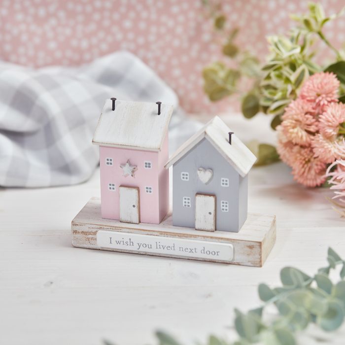 Two small wooden house figurines sit on a base with the inscription "I wish you lived next door." One house is painted pink with a star-shaped window, and the other is gray with a heart-shaped window. This I Wish You Lived Next Door Double House Ornament features flowers, greenery, and a gray and white cloth in the background.
