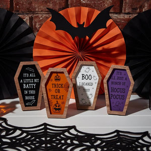 The image showcases high-quality wood Halloween Coffin Shaped Signs, perfect for spooky Halloween decor. Each sign features phrases like "It's all a little bit batty in this house," "Trick or treat," "Boo I scared you!" and "It's all just a bunch of hocus pocus." Behind is a paper bat and orange fan.