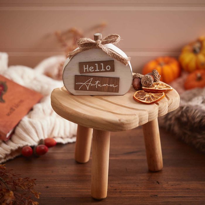 A small wooden stool holds a White Hello Autumn 3D Pumpkin Block with Bow, featuring the words "Hello Autumn." The stool is adorned with dried orange slices, acorns, and surrounded by cozy autumn decor including pumpkins and a knit blanket.