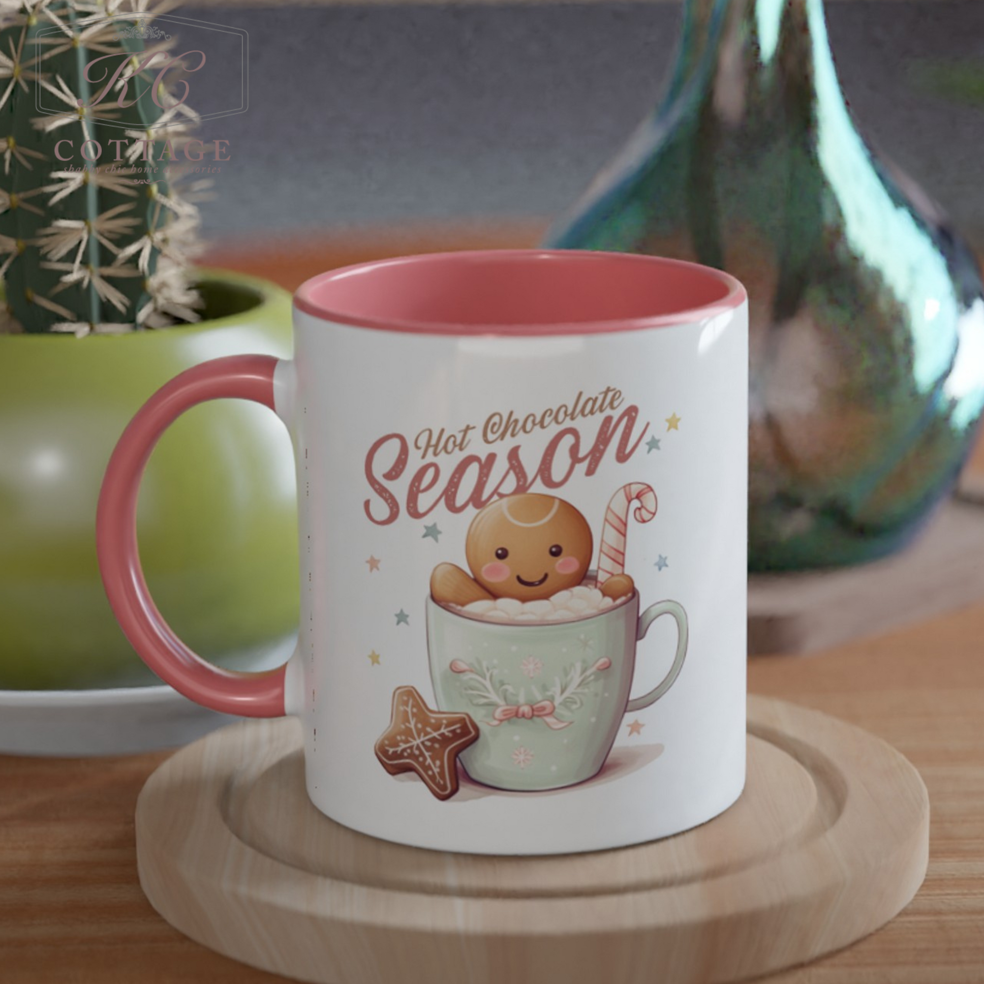 The Hot Chocolate Season Gingerbread Mug is a white ceramic 11oz mug with a pink interior, showcasing a design of a smiling gingerbread cookie sitting in a cup of hot chocolate. A candy cane and star-shaped cookie are also included in the scene, with "Hot Chocolate Season" text prominently displayed. In the background, you can spot a cactus and green vase.