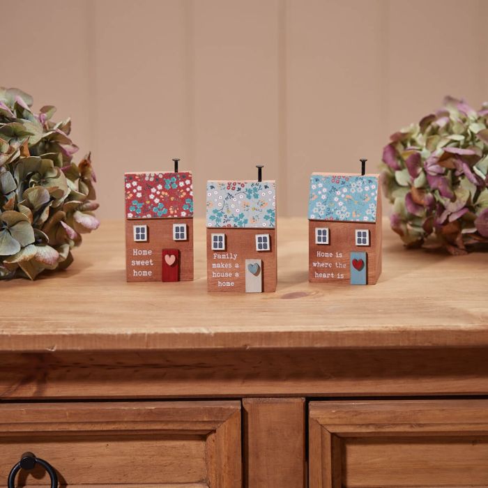 Three Wooden Houses With Floral Roofs and tiny chimneys are displayed on a wooden table. Each house features heartfelt sayings such as "Home sweet home," "Family makes a house a home," and "Home is where the heart is." Two clusters of hydrangea flowers sit nearby.