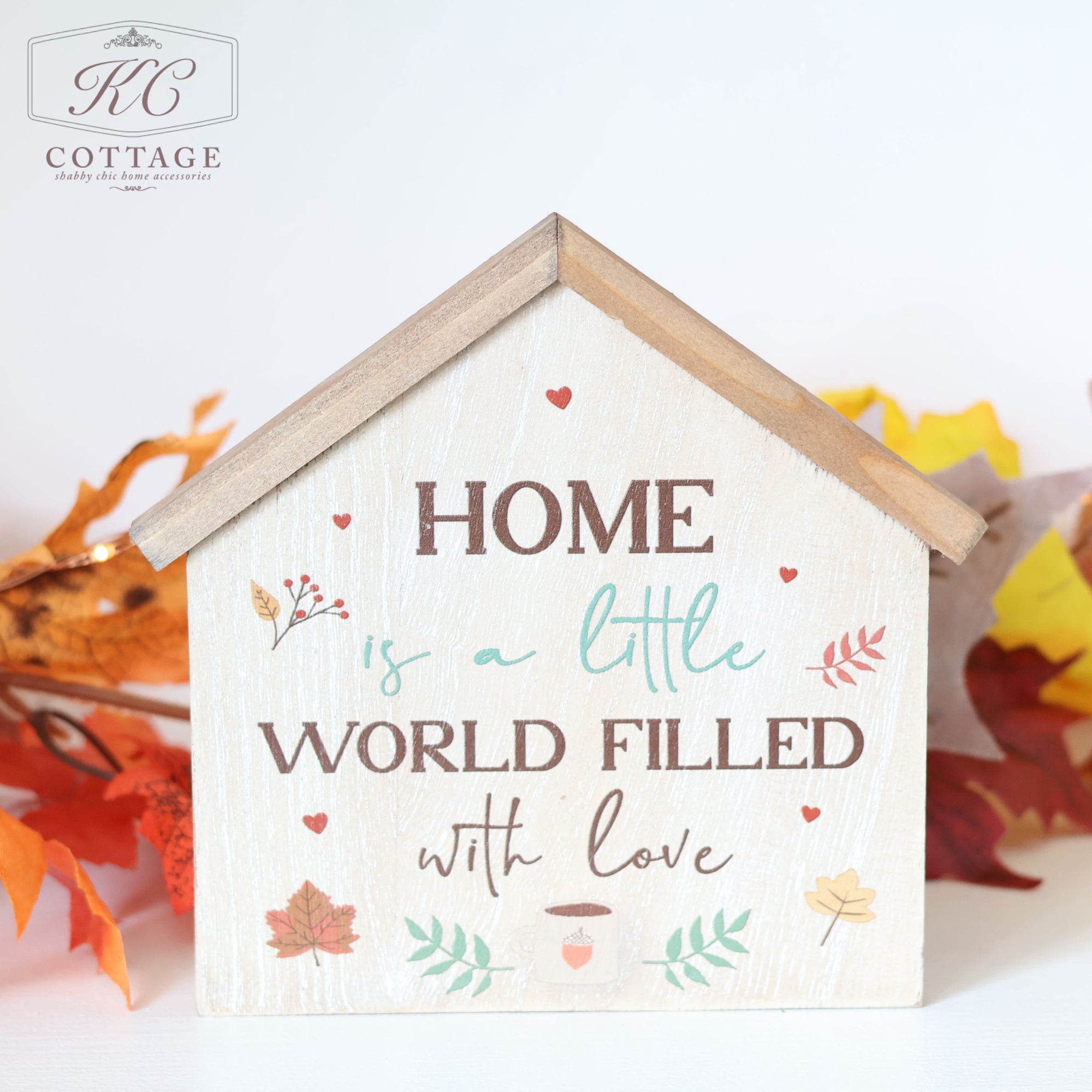 The Autumn Cosy Fall House Shaped Sign is a small, wooden decor piece in the shape of a house, featuring the text "Home is a little world filled with love." It is adorned with charming heart and leaf illustrations, with autumn leaves in warm colors completing the seasonal design. The "KC Cottage" logo appears in the top left corner.