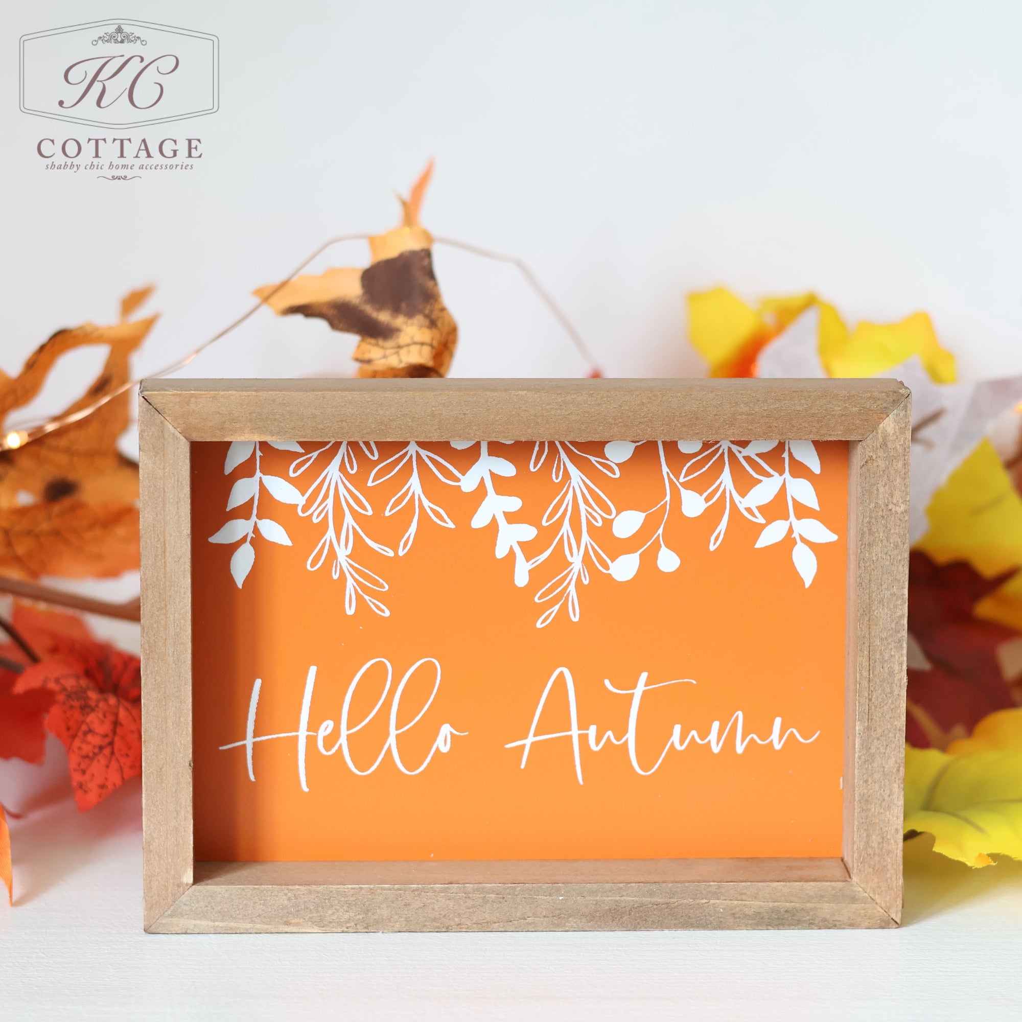 The "Autumn Pumpkin Patch Signs" feature a freestanding wooden-framed sign with an orange background, displaying the words "Hello Autumn" in elegant white cursive text. Above the greeting is a charming design of white leaves and branches. The sign is beautifully surrounded by colorful autumn leaves in shades of yellow, red, and orange, embodying the essence of Autumn Decor flawlessly.