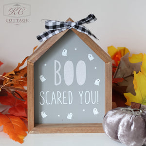 The Halloween Wooden House Sign features a gray background with the spooktacular phrase "BOO SCARED YOU!" surrounded by small white ghosts. Adorned with a black and white checkered ribbon, this haunted house style sign is set among autumn leaves and a pumpkin decoration.