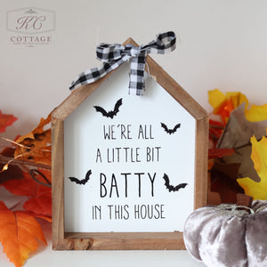 The Halloween Wooden House Sign, shaped like a house, showcases the spooktacular phrase "WE'RE ALL A LITTLE BIT BATTY IN THIS HOUSE" in black letters surrounded by bat silhouettes. It is topped with a black and white checkered ribbon and features a background adorned with autumn leaves and a decorative silver pumpkin.