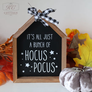 The Halloween Wooden House Sign, shaped like a small wooden house with a blackboard front, displays the text "It's all just a bunch of Hocus Pocus," surrounded by white stars. It is adorned with a black and white checkered ribbon, and is accentuated by autumn leaves and a silver fabric pumpkin.