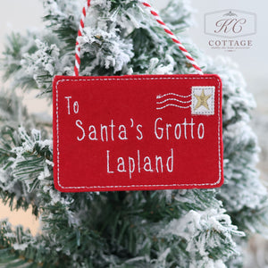 A red felt Christmas Hanging Santa Envelope adorns a snow-dusted tree, featuring the words, "To Santa's Grotto, Lapland," along with a gold star stamp design. This charming decoration captures the enchantment of sending a letter to Santa as a festive tradition.