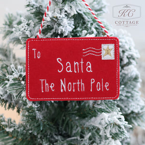 A red felt ornament, designed as a charming Christmas envelope with white stitching, hangs on a snowy Christmas tree. The sign reads "To Santa, The North Pole" and includes a gold star stamp design in the corner, perfectly capturing the festive spirit. This delightful decoration is known as "Christmas Hanging Santa Envelopes.