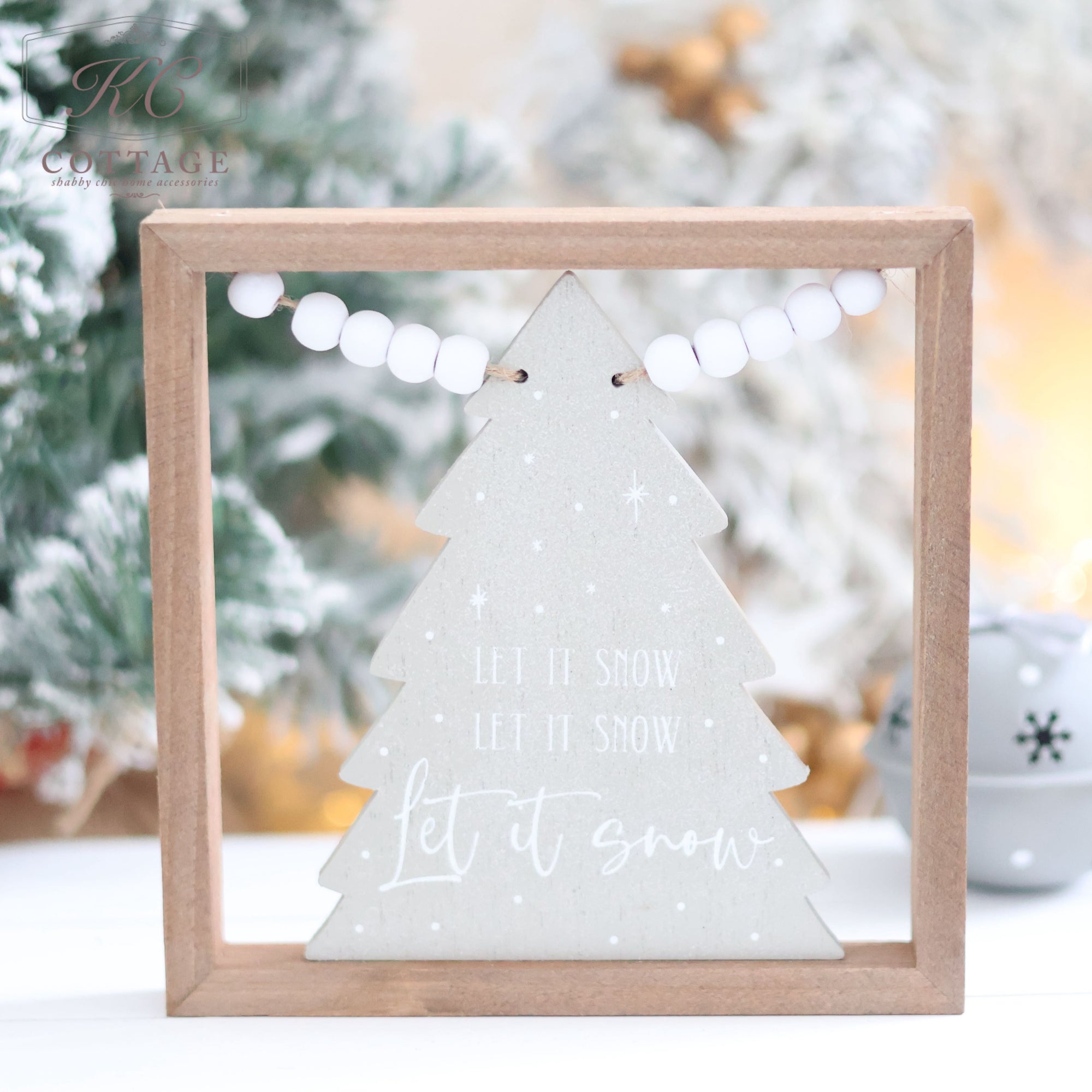 A Christmas Tree Sign with Beads features a wooden frame that highlights a light green tree board adorned with bead tinsel and the words "Let it snow" in white, set against a beautifully blurred snowy winter scene.