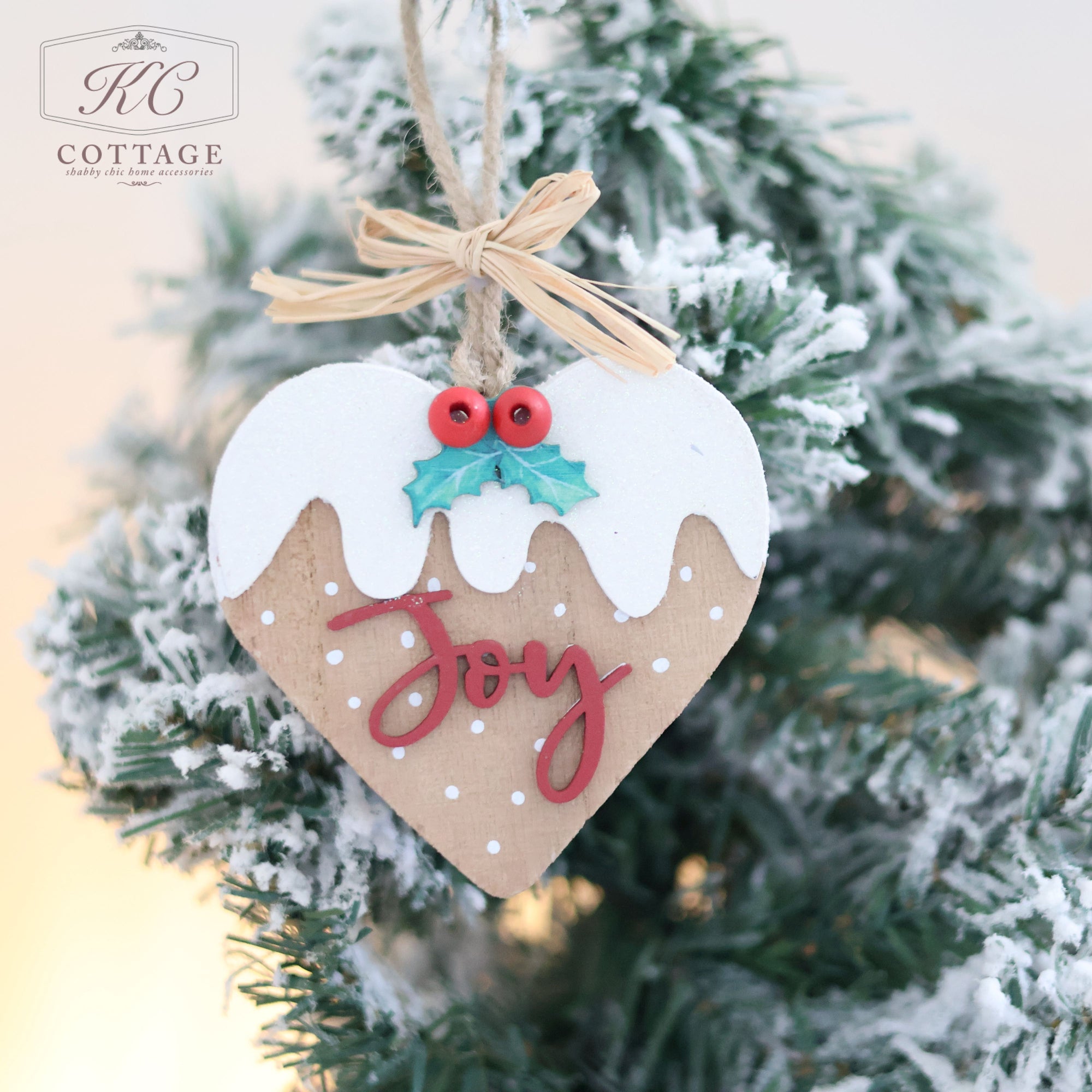 A charming Christmas Pudding Hanging Heart ornament, designed as a wooden heart and adorned with snow and holly, showcases the word "Joy" in vivid red as it elegantly hangs on a snowy branch.