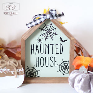 Introducing the Halloween Wooden House Sign, adorned with "Haunted House" text and detailed with spider web illustrations, a spider, and a bat. This decorative piece is enhanced by surrounding fall-themed accents like fabric pumpkins and autumn leaves. The KC Cottage logo in the top left corner adds a spooktacular finishing touch.