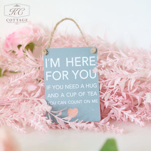 A small gray sign with white text saying "I'M HERE FOR YOU. IF YOU NEED A HUG AND A CUP OF TEA YOU CAN COUNT ON ME," is hanging against a background of soft pink foliage. The Gift Set - Thinking Of You also features a small peach-colored heart at the bottom.