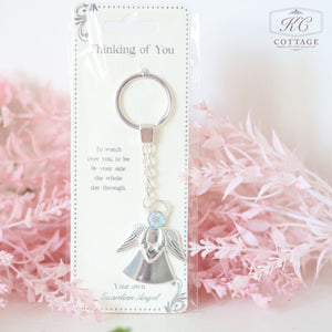 A silver keychain with an angel charm, featuring a small shimmering crystal, is attached to a card with the message "Thinking of You" and text about a guardian angel. The background has soft pink flowers, adding a touch of home decor elegance. This is part of the Gift Set - Thinking Of You.