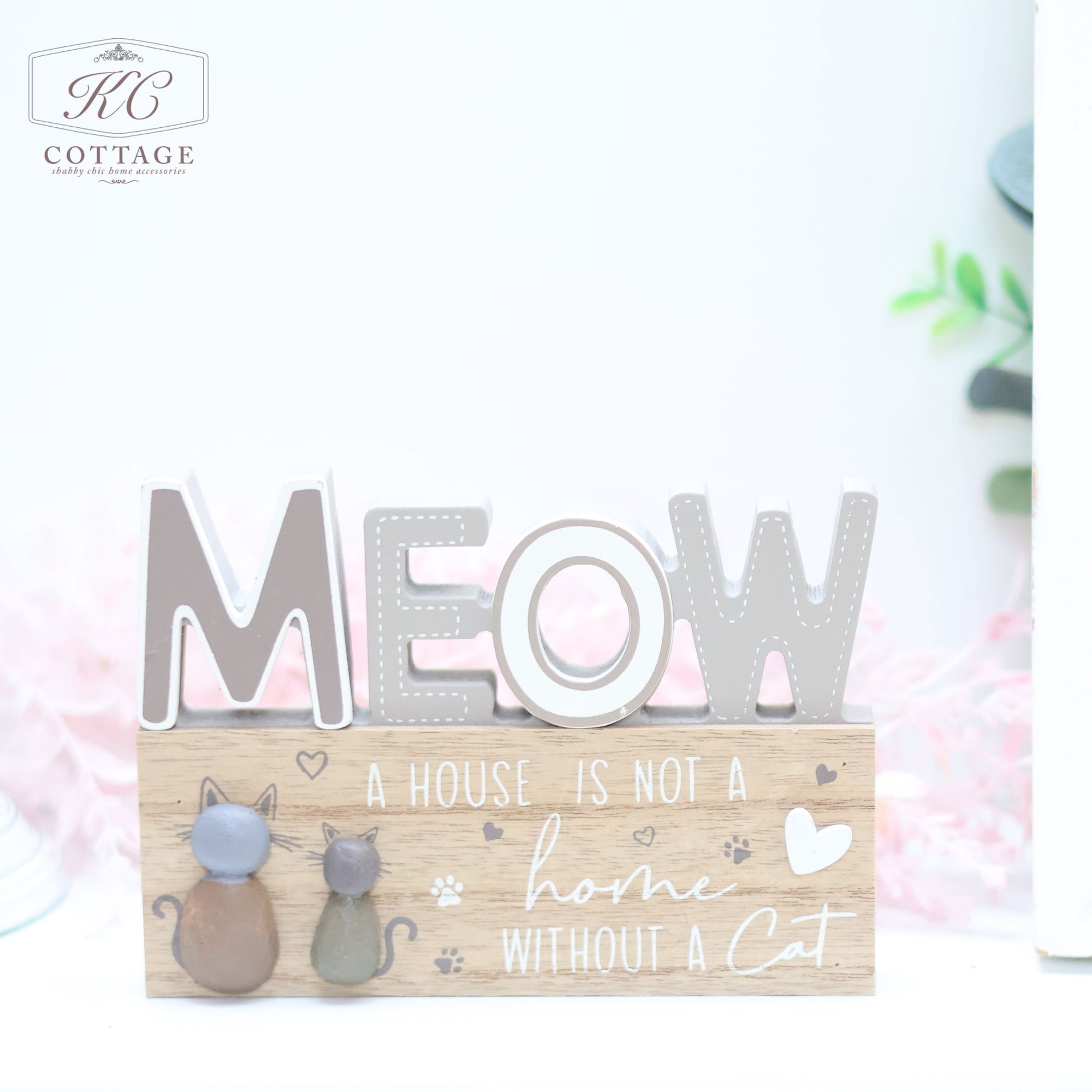 The Cat Pebble Signs features a charming decorative wooden block with the word "MEOW" prominently displayed on top. Below, it reads, "A HOUSE IS NOT A home WITHOUT A Cat." Two adorable cat figurines are affixed to the front. The background is softly blurred with hints of greenery, making it an ideal addition to any cat lover's home.