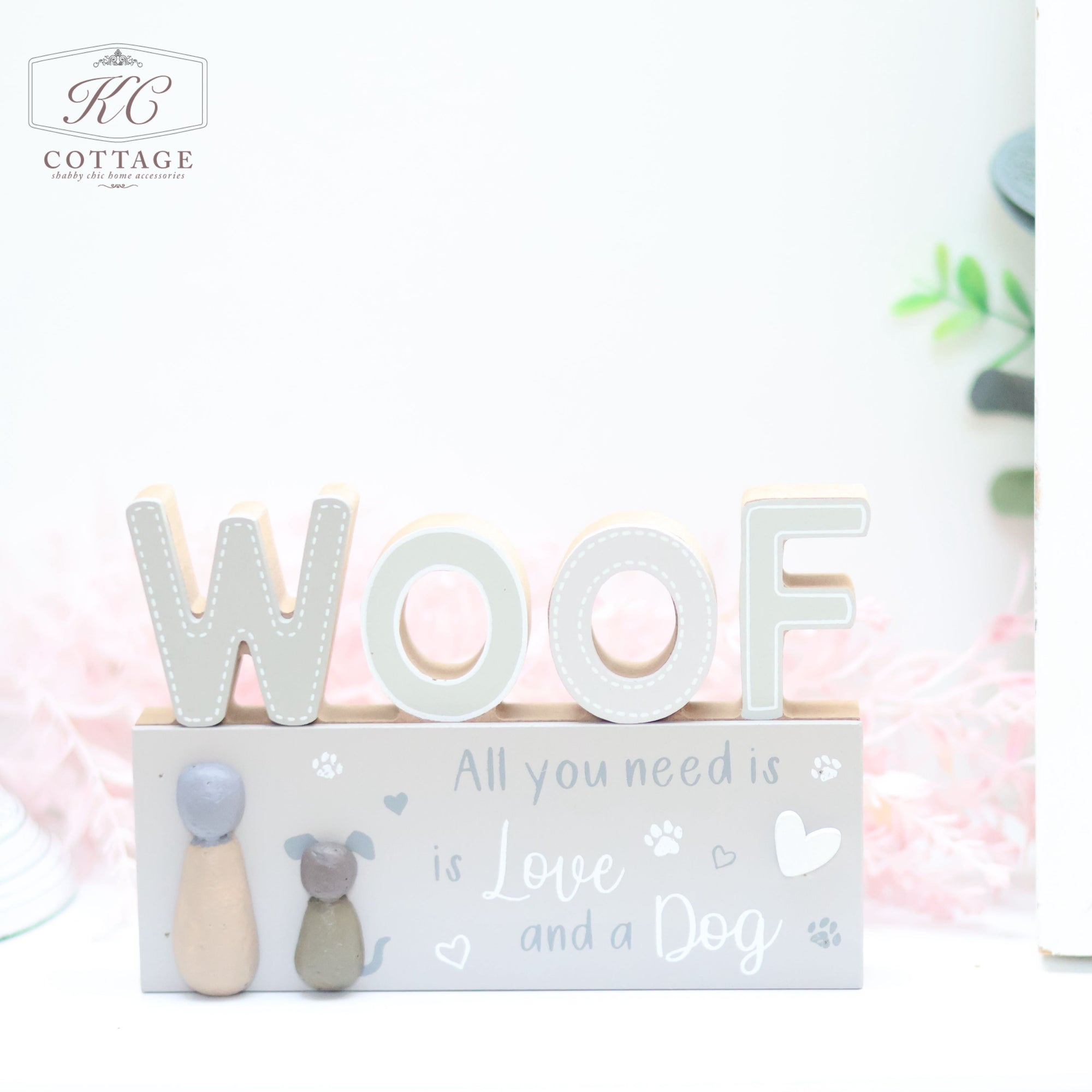 The Dog Pebble Signs, perfect for a dog lover's home décor, features large block letters spelling "WOOF" in white and beige. Below, it reads "All you need is Love and a Dog" with paw prints and heart motifs. To the left, cute figurines of a dog and a cat add charm. The upper left corner has a "KC Cottage" logo.