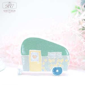 A small, whimsical Wooden Floral Caravan Ornaments shaped like a vintage camper with a green roof, yellow door, and floral accents. It sits against a soft, pastel background with hints of pink foliage. Perfect for adding charm to your home decor.
