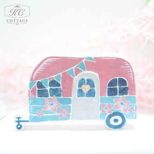 A whimsical illustration of Wooden Floral Caravan Ornaments, small, vintage-style camper trailers with red and teal colors adorned with floral and bunting decorations. The ornaments, perfect for home decor, have doors with heart-shaped windows and are displayed against a soft, white, and pink background.