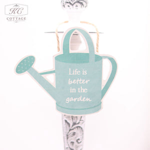 A Vegetable Garden Themed Hanging Sign shaped like a watering can, with the text "Life is better in the garden" written on it, hangs by a string from an ornately carved white post. The background is white, making the blue-green home decor sign and its message the focal point.