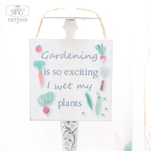 A Vegetable Garden Themed Hanging Sign, perfect for home decor, hangs by a rustic rope. It features illustrations of various vegetables and the playful text, "Gardening is so exciting I wet my plants." With a white background and a small "KC Cottage" logo in the top left corner, it adds charm to any space.