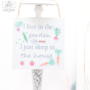 A charming piece of home decor, this Vegetable Garden Themed Hanging Sign hanging by a rope reads, "I live in the garden I just sleep in the house." Adorned with illustrations of tomatoes, radishes, carrots, peas, and greens around the text on a white background with a subtle weathered wood texture.