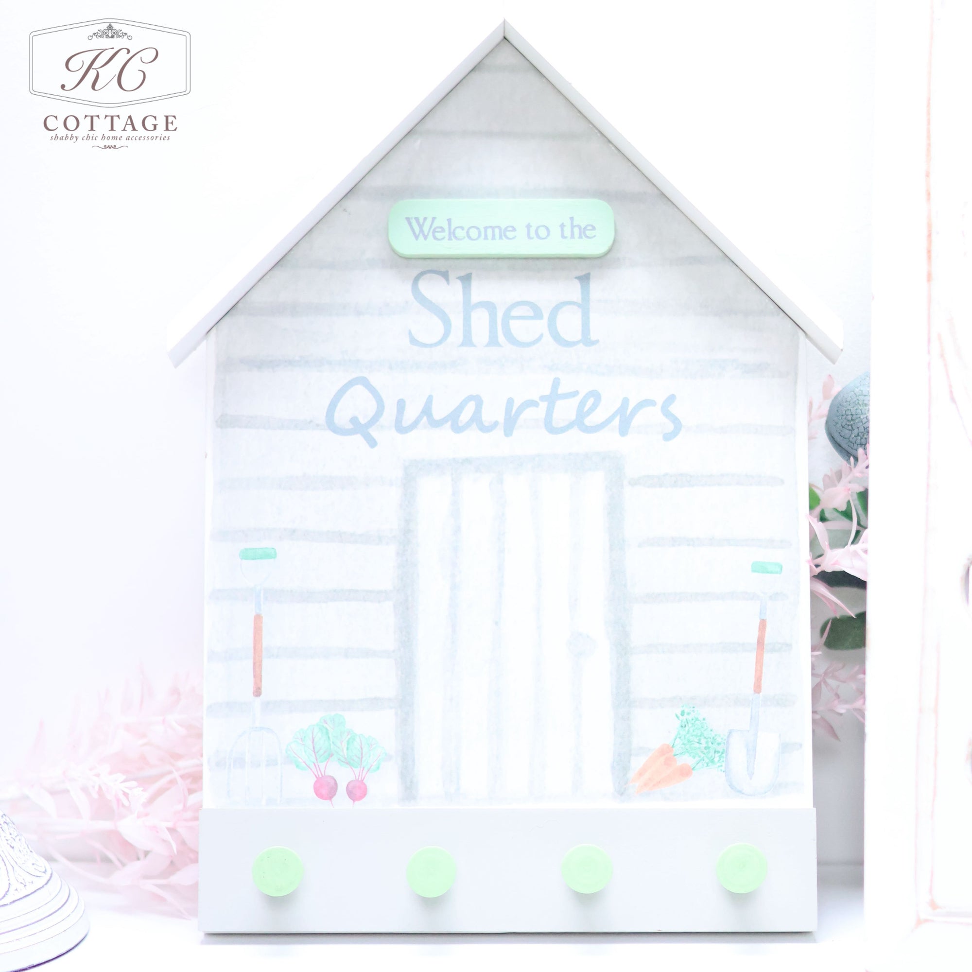 A Wooden Garden Shed Shaped Plaque with the phrase "Welcome to the Shed Quarters" written on the front. It features four green pegs at the bottom and a simple, light-colored design with small illustrations of gardening tools and vegetables—a charming piece of home decor.