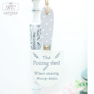 A small decorative Wooden Garden Trowel Hanger, perfect for home decor, hangs by a rope. The blade is white with "The Potting Shed - Where amazing things begin" written in gray. The handle is painted gray with white polka dots. In the background, a decorative candle holder and soft focus greenery add charm.