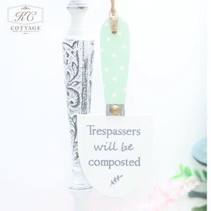 A Wooden Garden Trowel Hanger with a green and white polka-dotted handle hangs on a white, ornate stand. The trowel's blade has the humorous text "Trespassers will be composted" written in blue. Perfect for home decor enthusiasts, the display features a charming "KC Cottage" logo in the top left corner.
