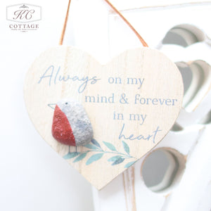 A Robin Pebble Hanging Heart with the text "Always on my mind & forever in my heart" and a small bird decoration in the lower left corner hangs from a string. Perfect for home decor, the sign rests against a white, heart-shaped cutout backdrop with a logo in the corner.