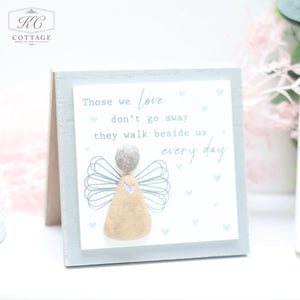 A charming piece of home decor, this Pebble Angel Forever Cards features an illustration of an angel made from pebbles and wire wings. The text reads: "Those we love don't go away, they walk beside us every day." Positioned on a light surface with a soft focus background adorned with pink flowers, it adds a heartfelt touch to any space.