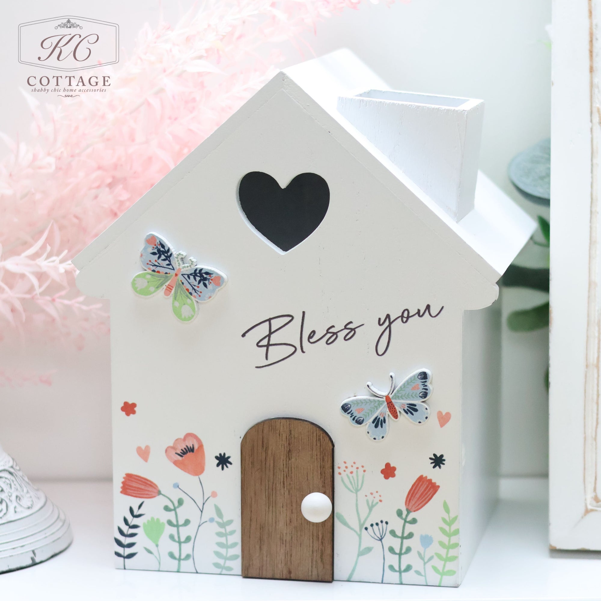 A Butterfly Tissue Holder House with a heart-shaped cutout at the top, adorned with butterfly and floral designs. The words "Bless you" are written on the front, making it perfect for home decor. The background features pink and green foliage, adding a charming touch.
