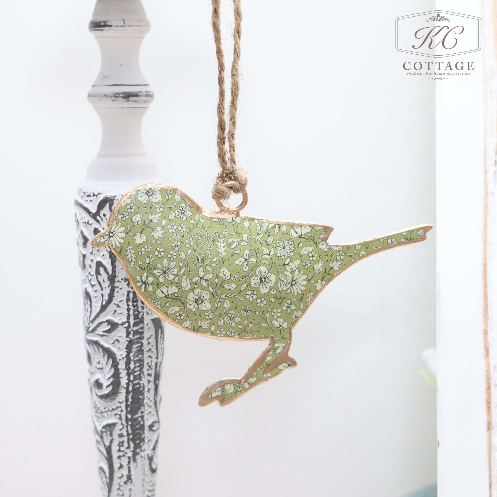 A Light Green Floral Metal Hanging Bird with a floral pattern hangs from a twine loop, adding charm to your home decor. Positioned next to a white and black intricately designed pillar, the bird features a green background with white flowers.