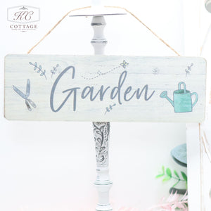 A rectangular wooden sign hanging from a rope reads "Garden" in cursive lettering, perfect for any nature lover. The sign is decorated with illustrations of gardening tools, a watering can, leaves, and small dotted lines. The light background enhances the rustic and charming aesthetic of these Wooden Garden Hanging Plaques.