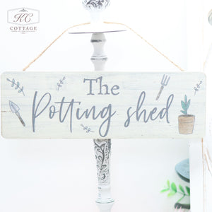 A rustic wooden garden sign with "The Potting Shed" written in cursive. Small illustrations of a trowel and fork adorn either side of the text, with a potted plant to the right. Perfect for the nature lover, these Wooden Garden Hanging Plaques hang beautifully by a piece of twine.