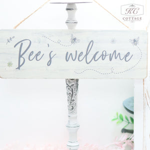 A white wooden sign with decorative floral elements reads "Bee's welcome" in elegant script. The Wooden Garden Hanging Plaques are displayed on a light-colored, intricately carved stand in a garden setting. The background is bright and simple, suggesting a cozy cottage atmosphere perfect for any nature lover.