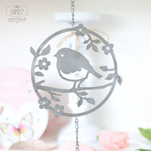 A Metal Robin Cutout Hanger Grey Metal With Bell featuring a silhouette of a bird on a branch adorned with flowers. The background is soft and blurred, showcasing a pastel-hued setting with floral elements, making it an exquisite piece of home decor.