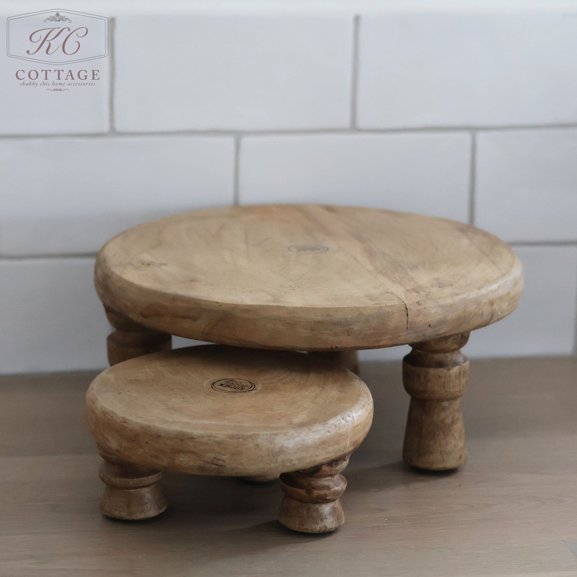 Two Raised Mango Wood Plates (set of two) of varying heights are placed on a wooden surface against a white tiled wall. Both plates have round surfaces and three legs each. The smaller plate is positioned slightly in front of and beneath the larger one, adding a charming touch to your home decor.
