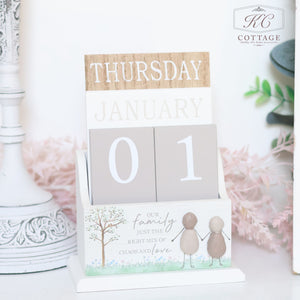 A decorative Pebble Family Calendar displays "Thursday, January 01" with cards and blocks in neutral tones. The base features a whimsical illustration of two stylized figures and the text, "Our family is just the right mix of chaos and love." It’s a perfect gift for a forgetful person.