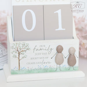 A wooden block calendar display with the numbers "01". The stand features an illustration of two figures holding hands beside a tree and the words "Our family just the right mix of chaos and love." The frame has a delicate floral design at the bottom – a perfect gift for forgetful person or Pebble Family Calendar.